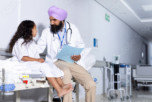Sikh male doctor in turban and biracial girl patient talking in corridor at hospital, copy space photo