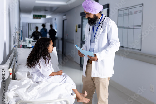 Happy sikh male doctor in turban and biracial girl patient talking in corridor at hospital photo