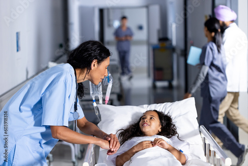 Biracial female doctor and smiling girl patient in bed talking in corridor at hospital photo