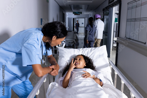 Biracial female doctor and smiling girl patient in bed talking in corridor at hospital photo