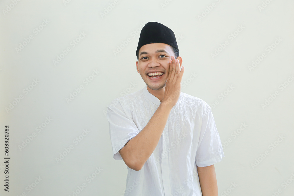Portrait young Asian Muslim man with hand near mouth whispering gossip and telling secrets while sharing news isolated over white background. People religious Islam lifestyle concept Portrait young Asian Muslim man with hand near mouth whispering gossip and telling secrets while sharing news isolated over white background. People religious Islam lifestyle concept