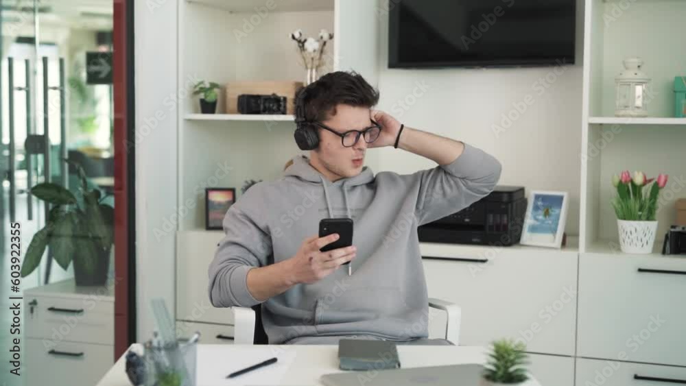 The very charismatic young man sits on a chair in front of the table and listens to music on wireless headphones. He holds a phone in his hand and switches tracks, enjoying the music. The very charismatic young man sits on a chair in front of the table and listens to music on wireless headphones. He holds a phone in his hand and switches tracks, enjoying the music.