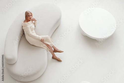 Horizontal from above view of elegant senior woman wearing fashionable dress sitting on white sofa in modern living room photo