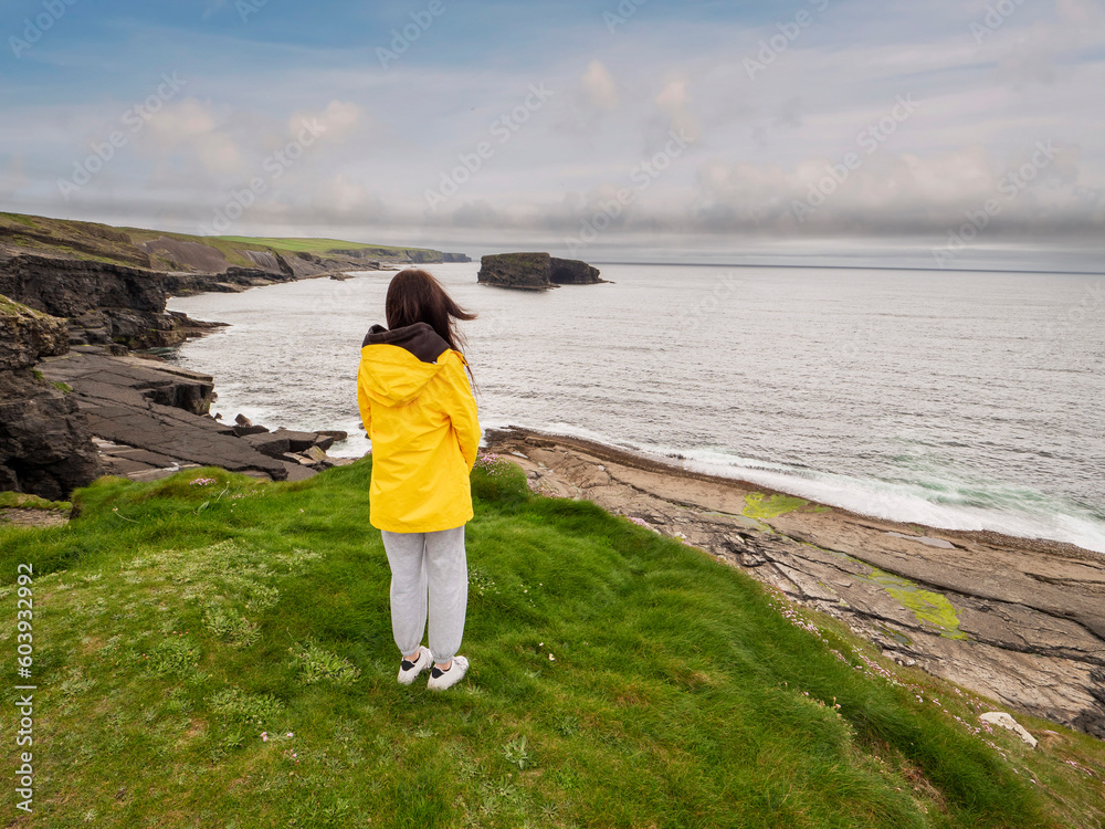 Teenager girl in yellow jacket looking at stunning nature scenery with cliffs and ocean, Kilkee area, county Clare, Ireland. Travel and sightseeing concept. Cloudy sky.