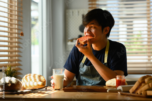 Fotografie Happy young asian man eating a fresh slice of bread with jam for breakfast while