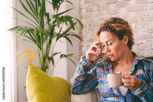 Thoughtful mature woman at home sitting on sofa with drink touching her temple head and looking down on the floor. Bad mind state condition. People worried and depression concept. Tired stressed lady