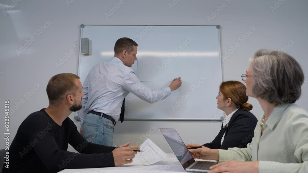 Caucasian man draws a graph on a white board, three colleagues sit at the table and listen to him.