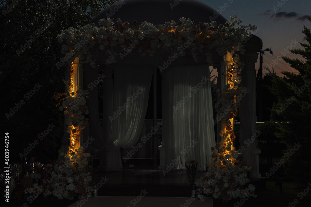 Wedding round arch decorated with cloth, flowers and lights outdoors in night. Wedding ceremony in the garden.	