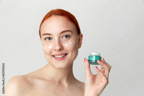 Cheerful beautiful young model, girl with bare shoulders and red hair showing, advertising eye cream over white background. Close Up