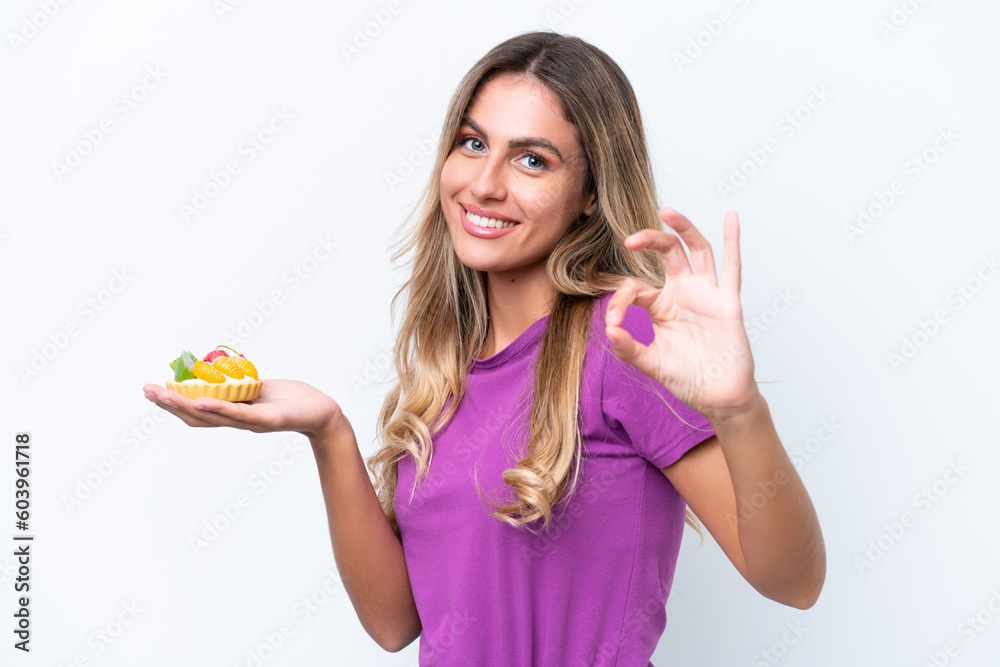 Young pretty Uruguayan woman holding a tartlet isolated on white background showing ok sign with fingers Young pretty Uruguayan woman holding a tartlet isolated on white background showing ok sign with fingers