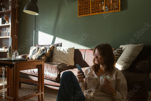 Woman sitting on floor by sofa with smart phone and cup