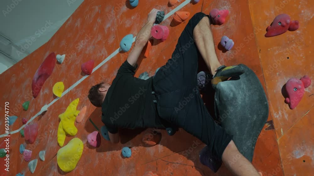 A professional athlete male millennial climber trains on a climbing wall with artificial stones indoors and performs a complex technique of rolling through the heel. High quality 4k footage