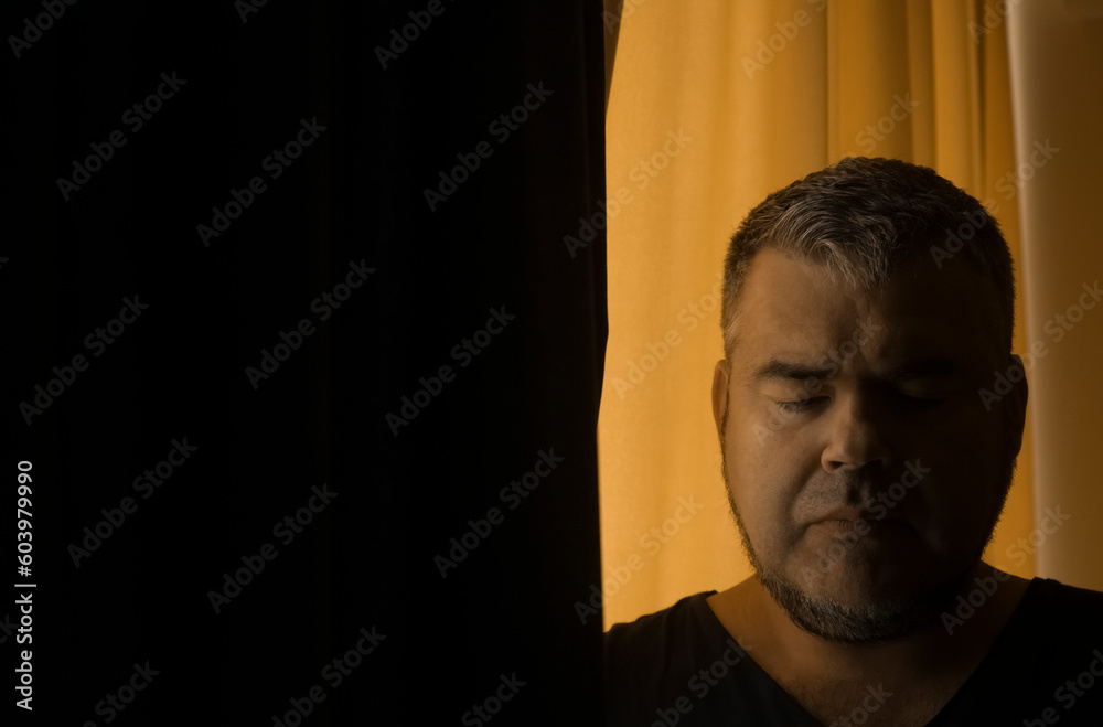 middle aged Caucasian man close up portrait with curtain. half of the face of male, guy is illuminated by light passing through yellow and black curtains. bearded guy looking down middle aged Caucasian man close up portrait with curtain. half of the face of male, guy is illuminated by light passing through yellow and black curtains. bearded guy looking down