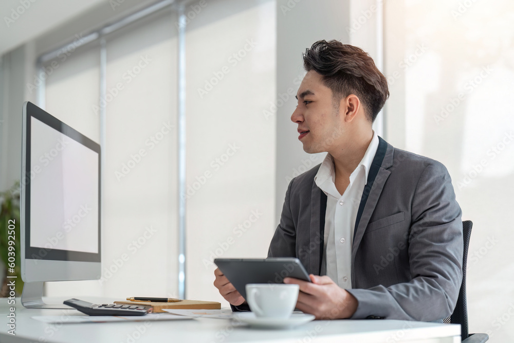 Businessman looking at computer monitor holding tablet sitting in office