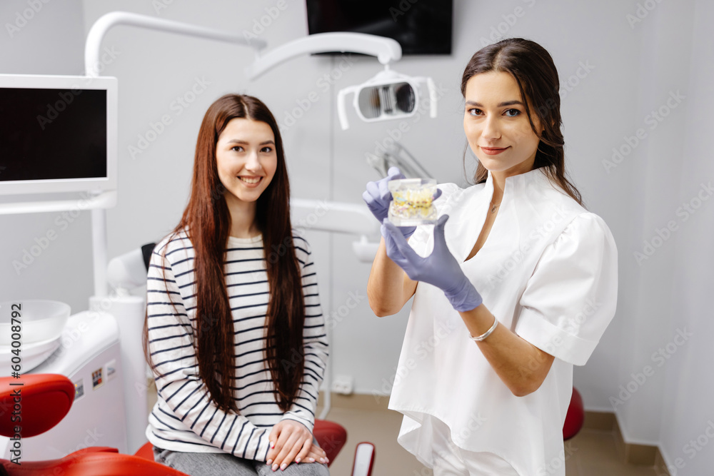 Young woman having a visit at the dentist. Female doctor showing fake plastic jaw. Beautiful girl patient sitting on chair at dentist office in dental clinic. Healthy teeth and medicine concept.