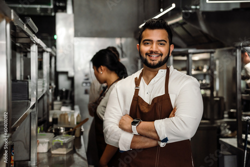 Positive busy indian male business owner in apron looking at camera in cafe kitchen