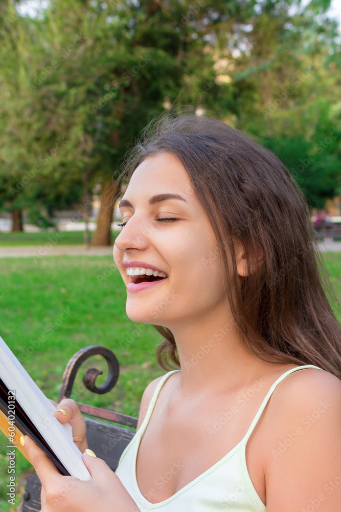 Fototapeta premium Beautiful young brownhair girl smells a fresh printed book sitting on the bench in the park