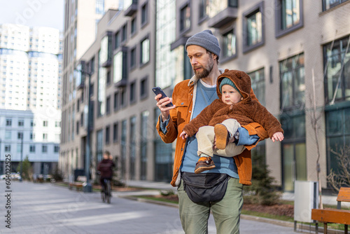 A man with a child on his back walks down the street, holding a phone and looking at the screen