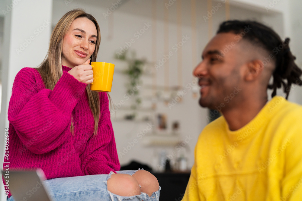 Smiling young girlfriend with long hair in pink sweater sitting on table and talking with Indian boyfriend while drinking tea from yellow mug Smiling young girlfriend with long hair in pink sweater sitting on table and talking with Indian boyfriend while drinking tea from yellow mug