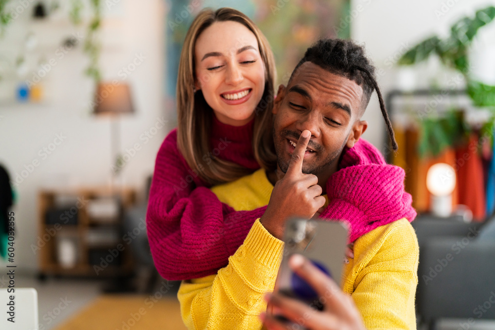 Delighted young multiracial couple in colorful wool sweater smiling and taking selfie on smartphone while sitting at wooden table together in modern apartment Delighted young multiracial couple in colorful wool sweater smiling and taking selfie on smartphone while sitting at wooden table together in modern apartment