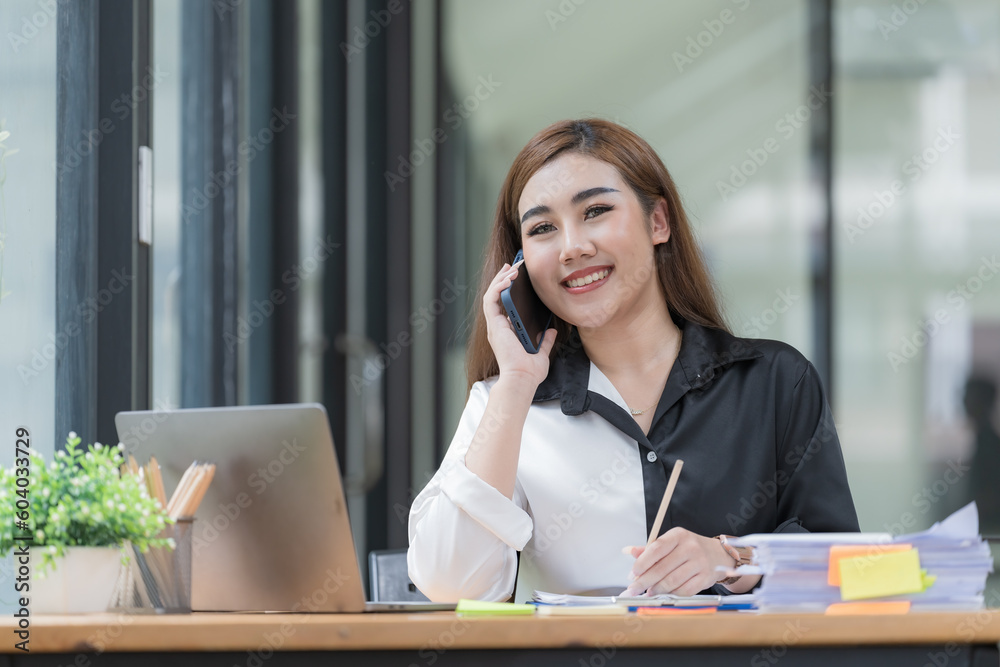Happy young Asian businesswoman talking on the mobile phone and smiling while sitting at her working place in the office. Happy young Asian businesswoman talking on the mobile phone and smiling while sitting at her working place in the office.