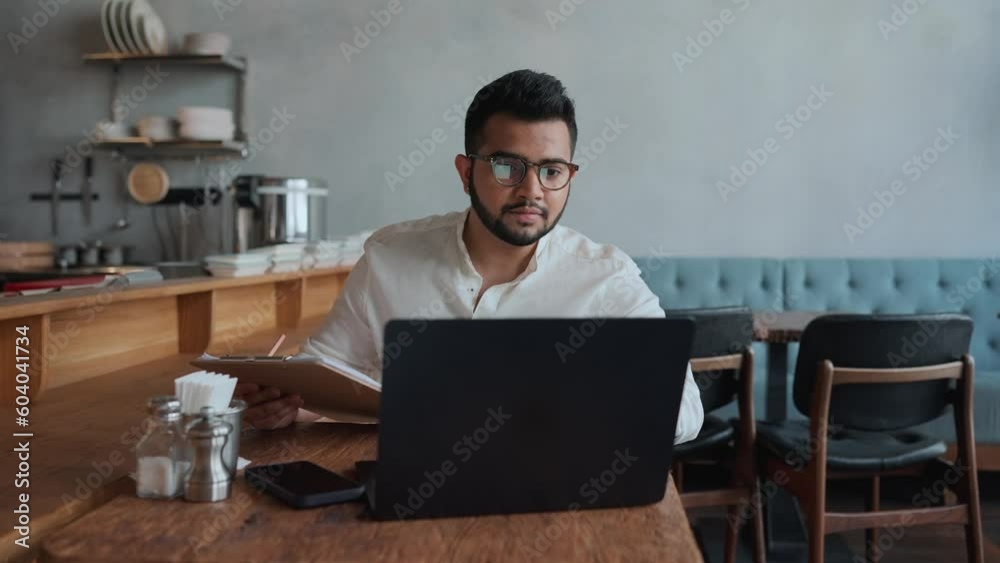 Serious indian male business owner in eyeglasses filling orders on laptop in cafe Serious indian male business owner in eyeglasses filling orders on laptop in cafe