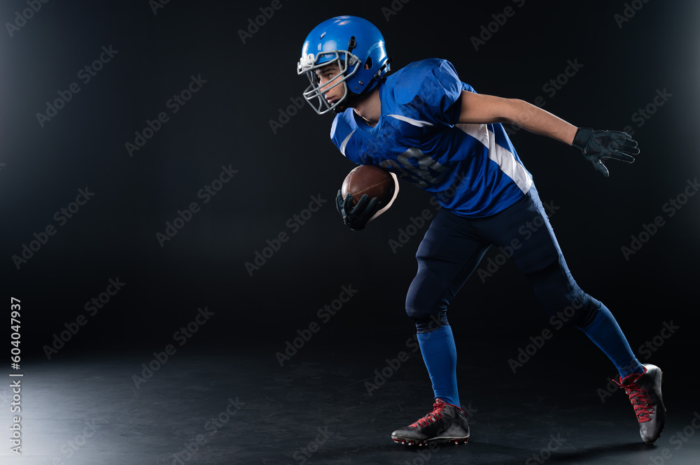 Full length portrait of a man in a blue american football uniform against a black background. Sportsman in a helmet with a ball. 