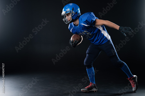 Full length portrait of a man in a blue american football uniform against a black background. Sportsman in a helmet with a ball. 