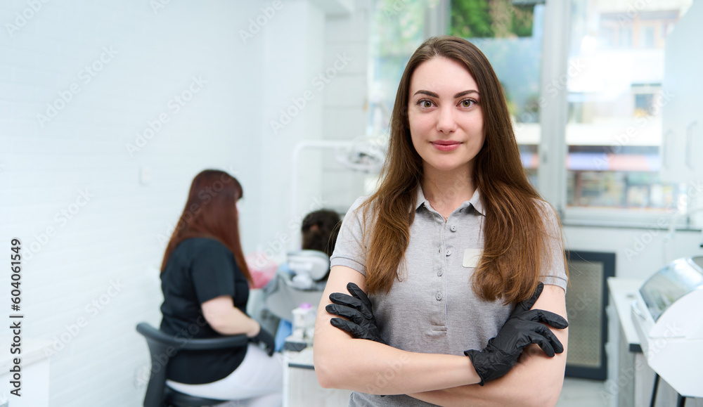 Obraz premium Confident portrait of a young female doctor dentist hygienist, healthcare worker, dental assistant posing with her arms folded, looking at camera, standing in her workplace in modern dentistry clinic
