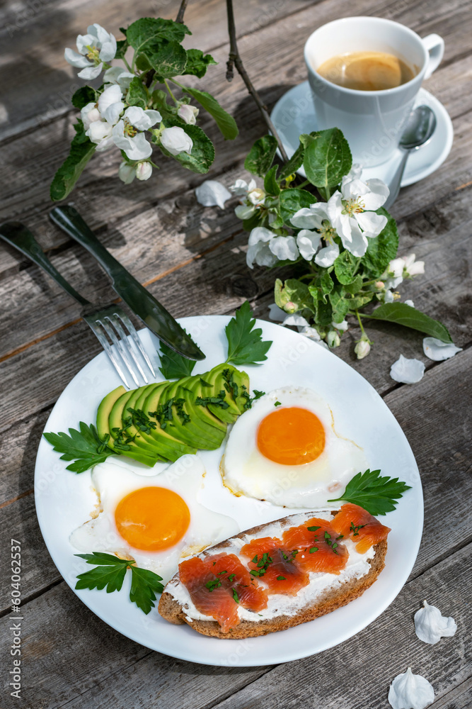 Sandwich with salted trout and avocado for breakfast. Two fried eggs on a plate. A cup of coffee on a wooden table. Sandwich with salted trout and avocado for breakfast. Two fried eggs on a plate. A cup of coffee on a wooden table.