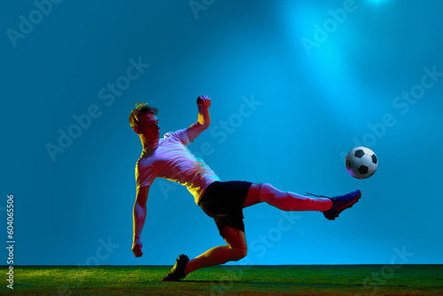 Kicking the ball. Professional football player in sports uniform in action over soccer field background in neon light. Concept of active life, team game