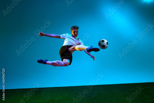 Professional man  football player in sports team uniform kicking the ball in motion over soccer field background in neon light. Scoring a goal.