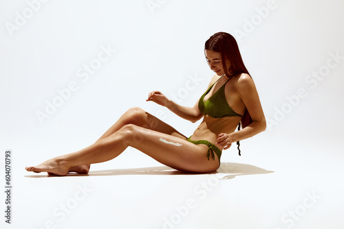 Summer protection. Portrait of beautiful young woman with tanned body in swimsuit, applying spf cream against white studio background. Concept of body, skin care, spf protection, cosmetics, beauty