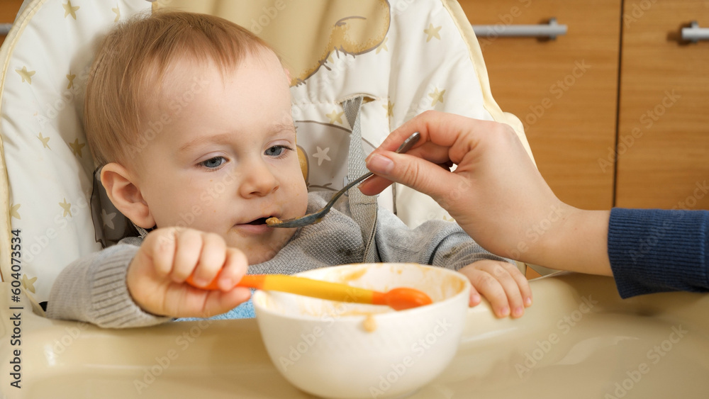 Closeup portrait of baby boy holding spoon while mother is feeding him in highchair. Concept of parenting, healthy nutrition and baby feeding. Closeup portrait of baby boy holding spoon while mother is feeding him in highchair. Concept of parenting, healthy nutrition and baby feeding.
