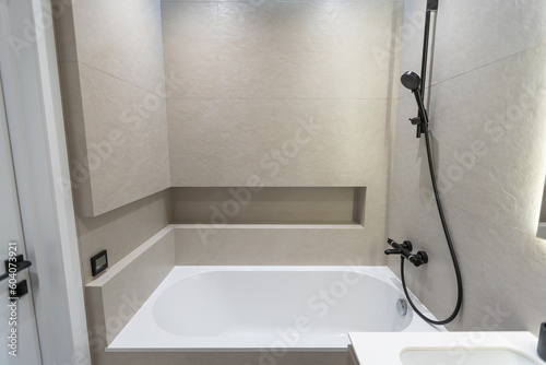 Bathroom in a modern style with gray and white tiles. There is a large mirror with luminous lamps, tabletop with wooden drawers and sink, bath with shower and glass partition, towel rack and a hanger.