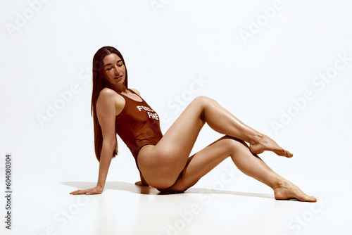 Portrait of beautiful young woman withslim, tanned body in swimsuit sitting against grey studio background. Concept of body, skin care, spf protection, cosmetics, beauty, summer
