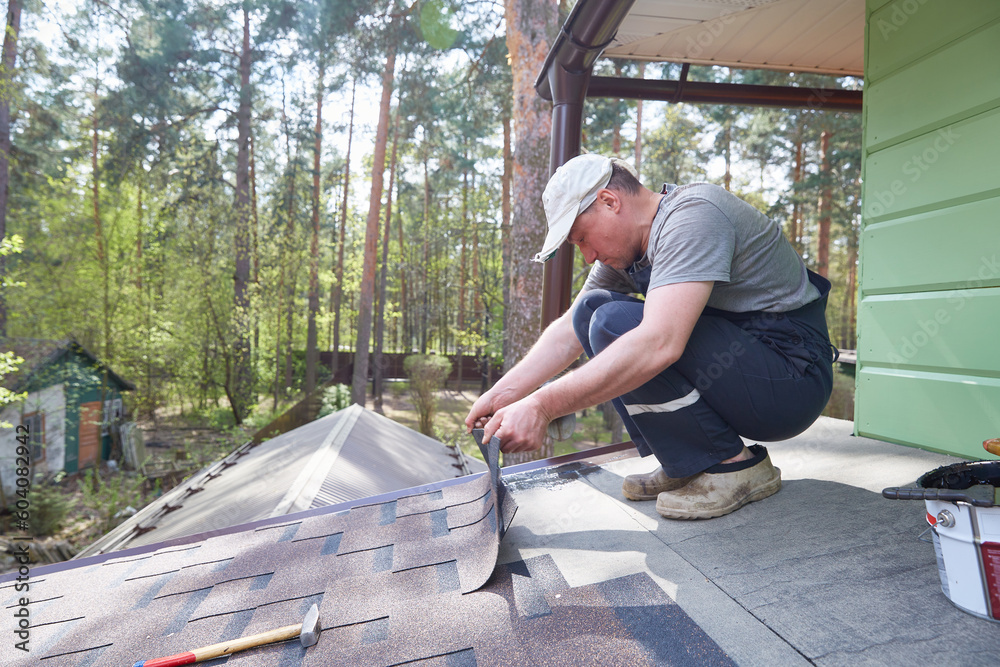 Male worker lays a sheet of soft flexible bitumen shingles on the roof of a country house. Male worker lays a sheet of soft flexible bitumen shingles on the roof of a country house.