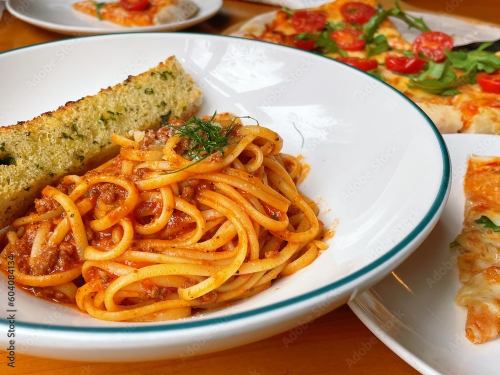Closeup shoot of spaghetti bolognese, pasta on a white plate, pizza as background on a table, western food Closeup shoot of spaghetti bolognese, pasta on a white plate, pizza as background on a table, western food