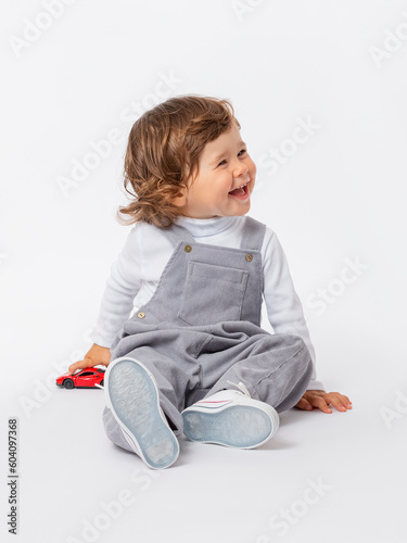 A cheerful laughing Caucasian toddler 2 years old with curly hair in a gray jumpsuit and a white turtleneck is playing with a car on the floor.