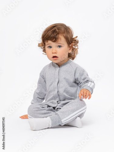 A sad 2-year-old toddler boy with curly hair in a gray jumpsuit and socks sits cross-legged on the floor and looks with an expressive look with his mouth open in surprise on a white background.
