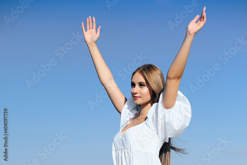 Happy, beautiful woman on the ocean beach standing in a white summer dress, raising hands.