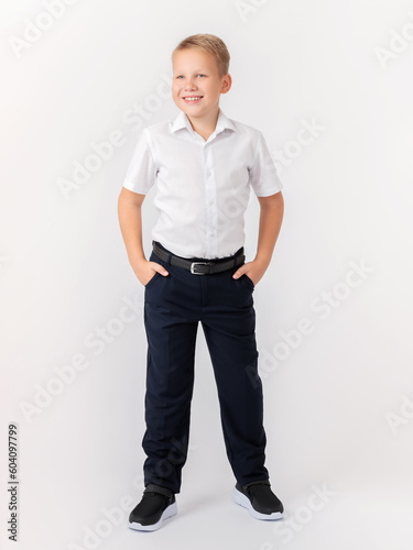 Cute confident teenager 10 years old blonde stands with his legs apart in a classic white shirt and dark trousers with a belt on a white background. Hands in pockets. Layout, advertising.