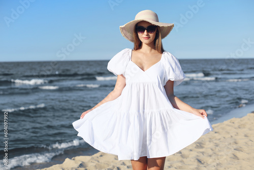 Happy smiling blonde woman is posing on the ocean beach with sunglasses and a hat.