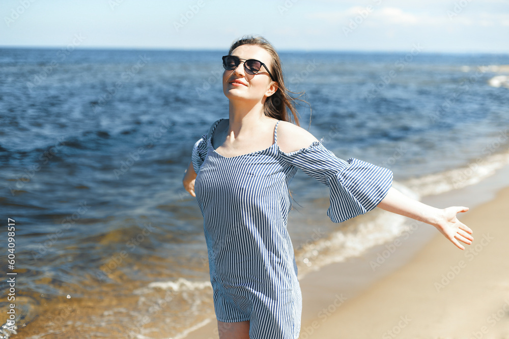 Happy smiling woman in free bliss on ocean beach standing with open hands. Portrait of a brunette female model in summer dress enjoying nature during travel holidays vacation outdoors