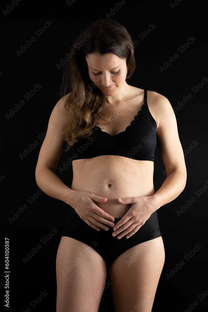 Obraz premium View of happy woman holding her belly gently during first months of pregnancy. Pregnancy first trimester - week 18. Front view. Black background. Bright shot.