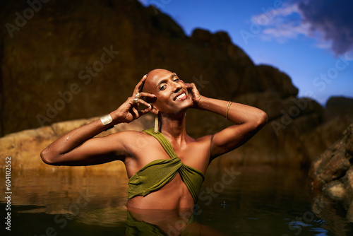 Happy trans sexual fashion model shows golden jewelry chest deep inside natural pool surrouded by rocks at night. Androgynous ethnic person stands in the middle of a backwater exotic scenic location photo