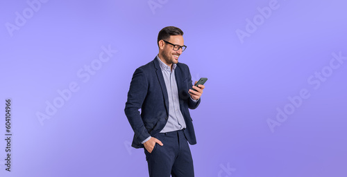 Male business professional dressed in elegant formals smiling and messaging online over mobile phone. Handsome chief with hand in pocket using cellphone while posing against blue background