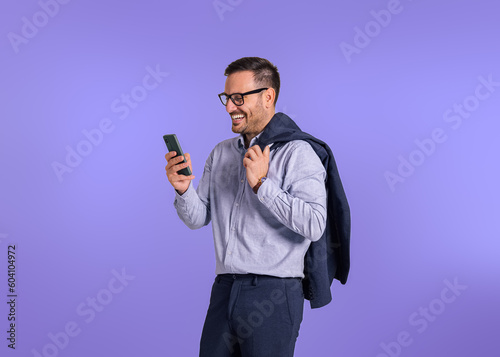 Happy young businessman holding blazer and checking messages on smart phone. Male professional entrepreneur smiling and scrolling social media while standing isolated against blue background © Moon Safari