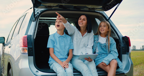 Family mother son and daughter countryside and talking while sitting in vehicle trunk in sunset. Discussing the right direction they will go with parents on vacation.