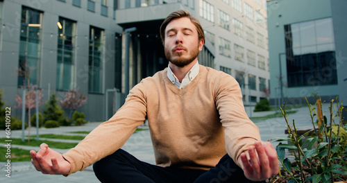 Handsome caucasian bearded man in glasses relaxing, sitting in the lotus position on the bench of the courtyard of office building or university. Healthy lifestyle, outdoors